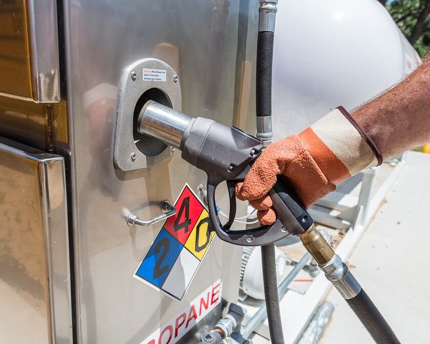 Gloved hand holding a fuel delivery nozzle connected to a hose and inserted into a dispenser. 