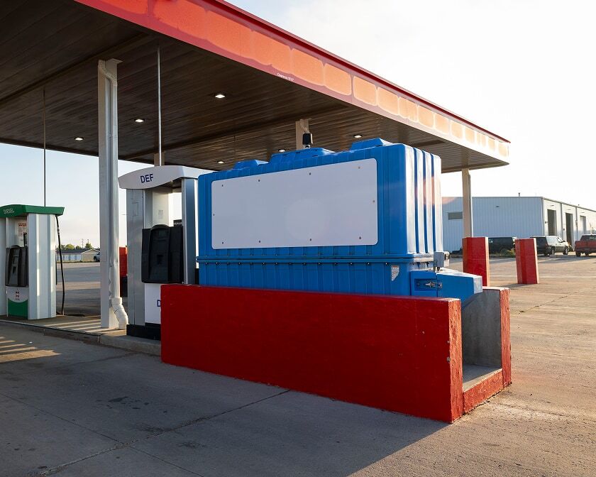 Blue DEF storage tank and dispenser under a fuel station canopy, with pump islands and red protective barriers around the tank.