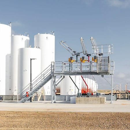 Fuel terminal loading rack with elevated platform and loading arms, multiple white vertical storage tanks, and a fuel delivery truck on the concrete pad.fuel-terminal