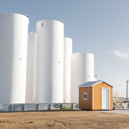 Industrial bulk plant with multiple large white vertical storage tanks, a small utility building, and a loading rack platform under a clear sky.