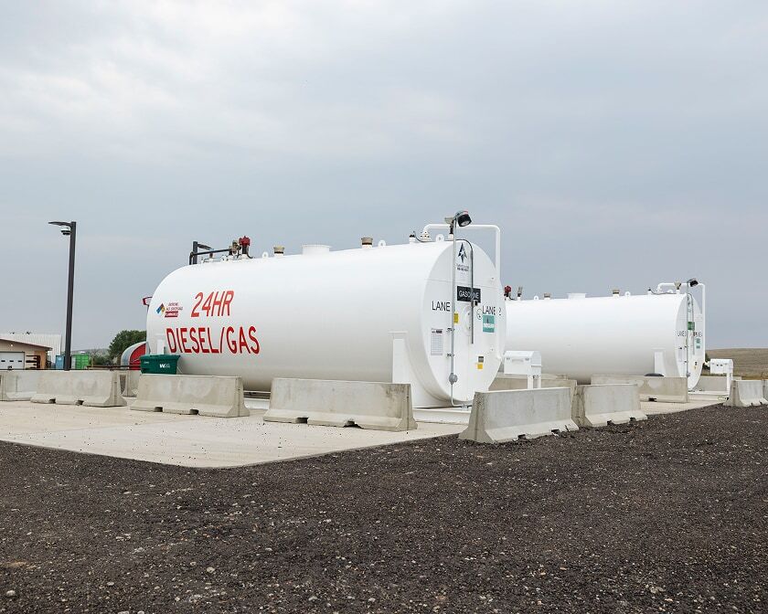 Two large white aboveground fuel tanks on a concrete pad with protective barriers at an outdoor fueling site.