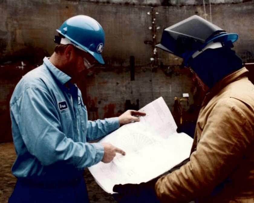Two workers wearing hard hats review a large blueprint inside an industrial facility.