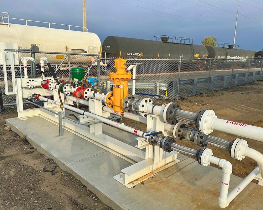 Propane metering skid with white piping, valves, and hoses mounted on a concrete pad inside a fenced facility, with railcars in the background.
