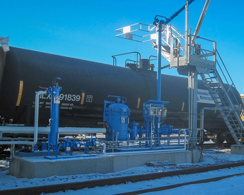 Rail loading skid with blue piping, valves, and control equipment beside a black tank railcar, with a loading platform and stairs at a rail terminal.