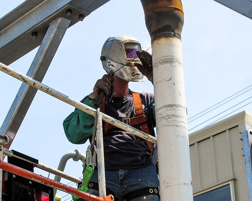 Technician wearing welding PPE and fall protection welds a pipe on an elevated work platform at a terminal. 
