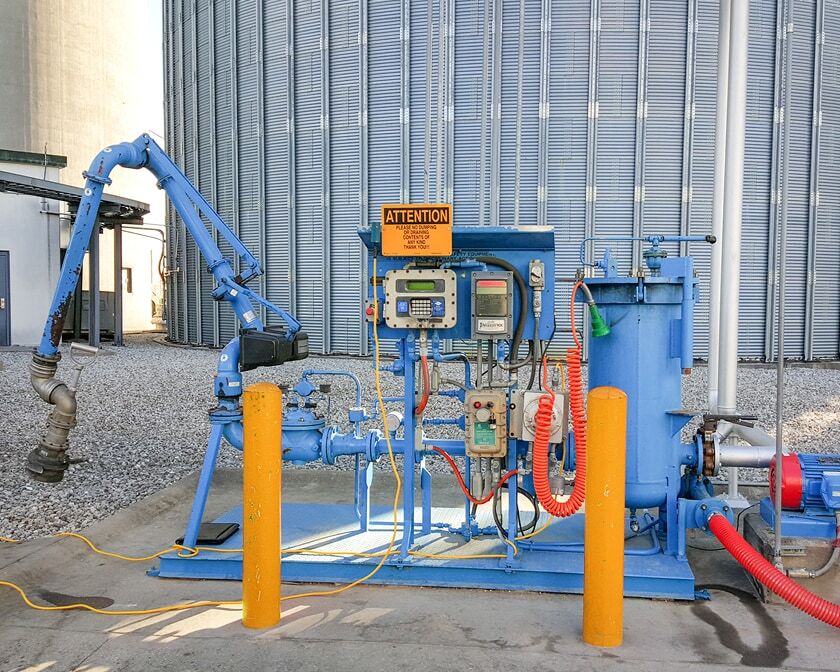 Blue truck loading skid with control panel, piping, and loading arm protected by yellow bollards at a terminal site in front of a large storage tank.