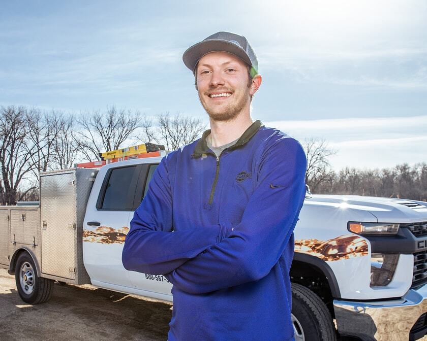 Smiling field service technician standing with arms crossed in front of a white service truck parked outdoors.